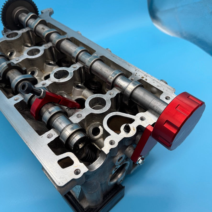 Red anodized cam bridge tool installed on a VW/Audi 2.0T cylinder head, hook arm clamping the camshaft.