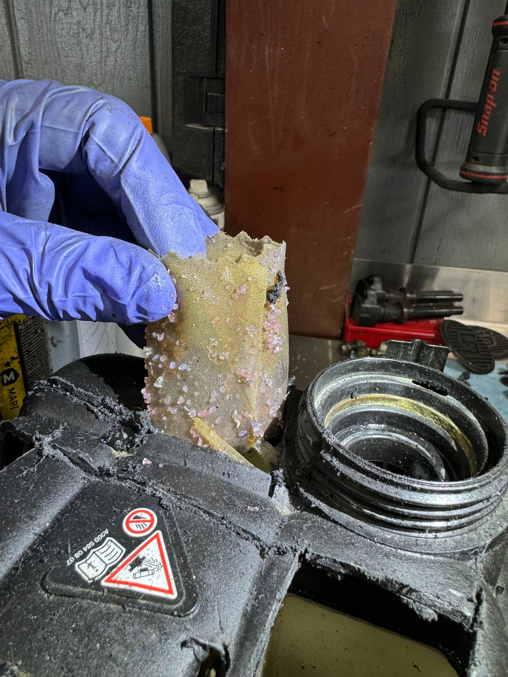 Gloved hand pulling a ruptured, crystal-caked filter bag from a failed Mercedes coolant housing.