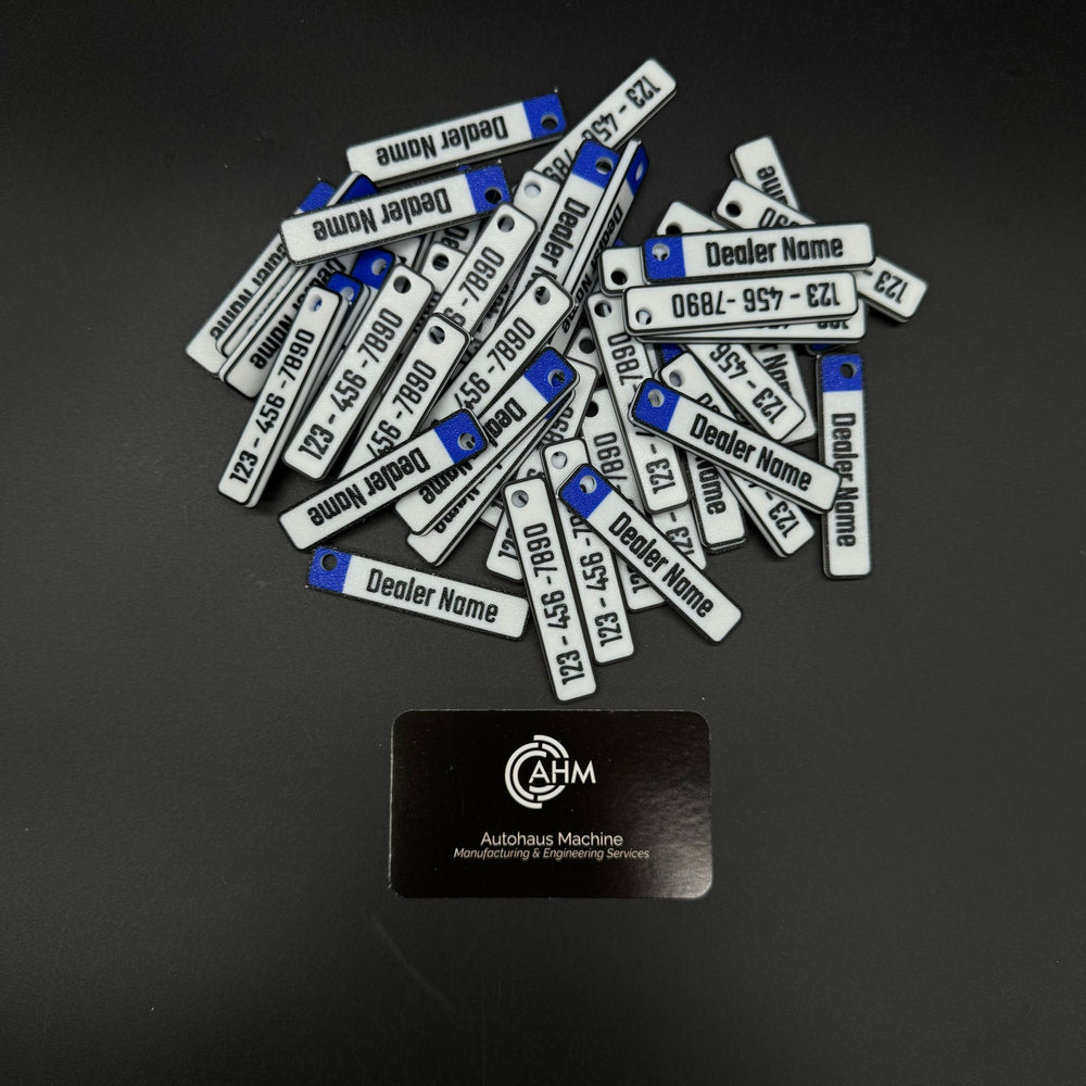 Pile of custom euro-plate dealership keychains, white with blue tab, engraved dealer name and phone.