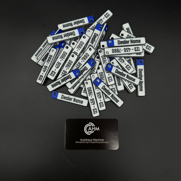 Pile of custom euro-plate dealership keychains, white with blue tab, engraved dealer name and phone.